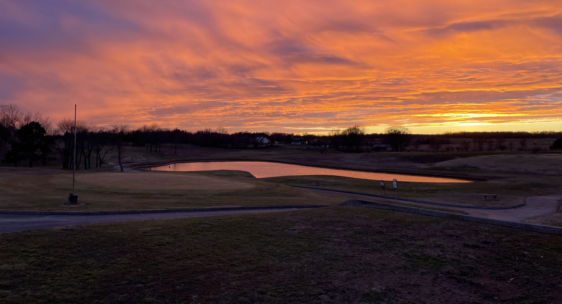 Sunset over a golf course with a pond reflecting vibrant orange and purple skies.
