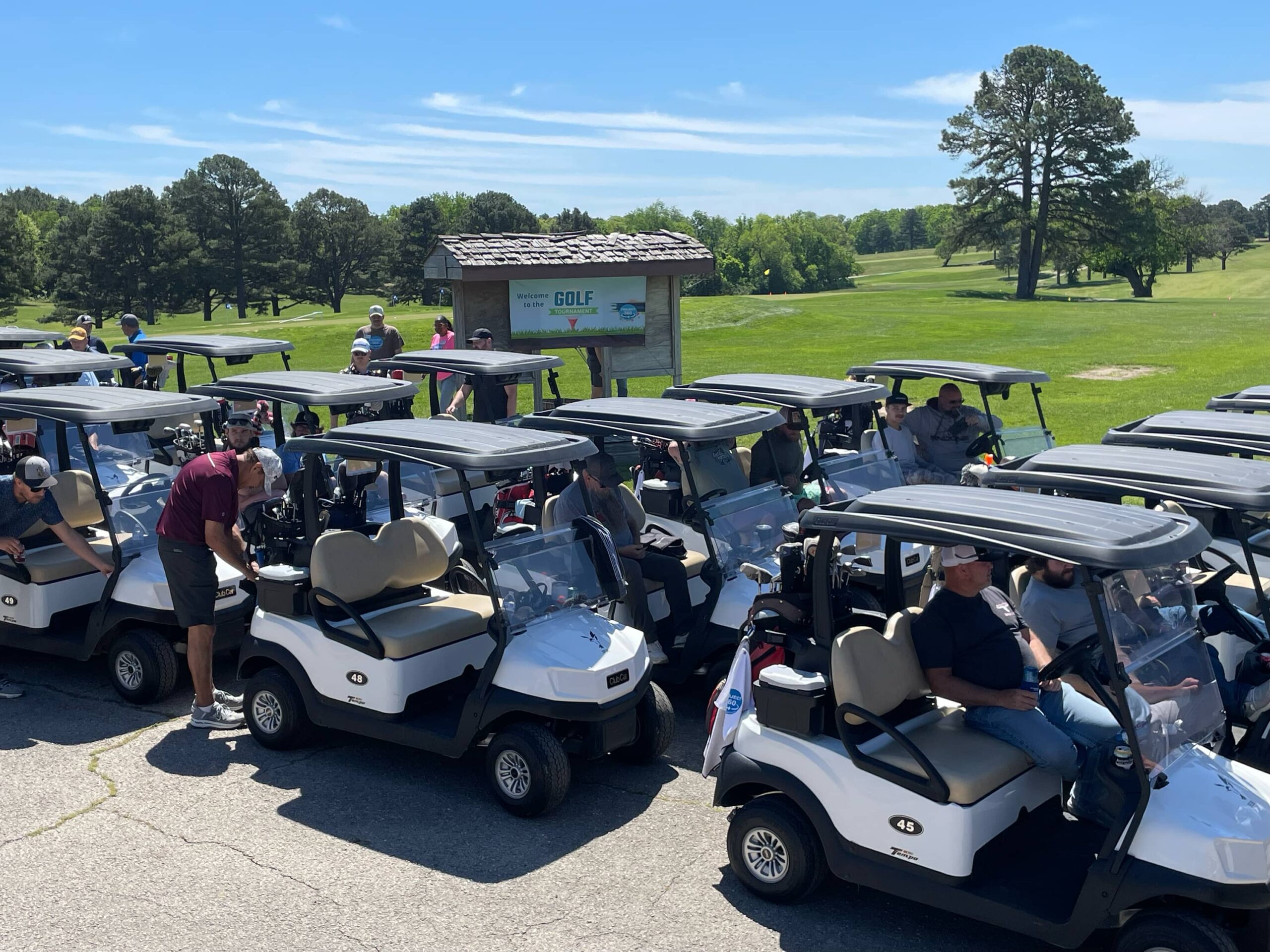 Golfers preparing to start a tournament, with multiple golf carts lined up and a green course in the background.